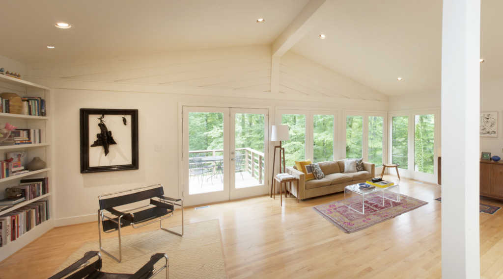 A panoramic view of the bright open living space with cathedral ceilings, built-in book shelves, warm wood floors and large windows