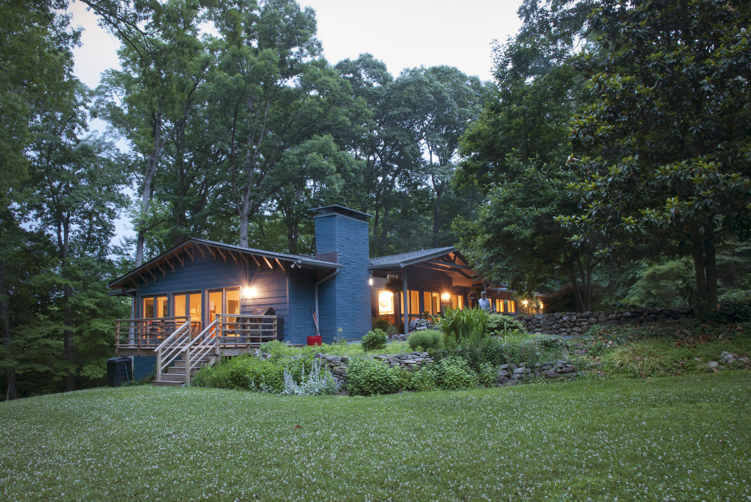 The house glows from within at dusk, surrounded by a lush garden and trees