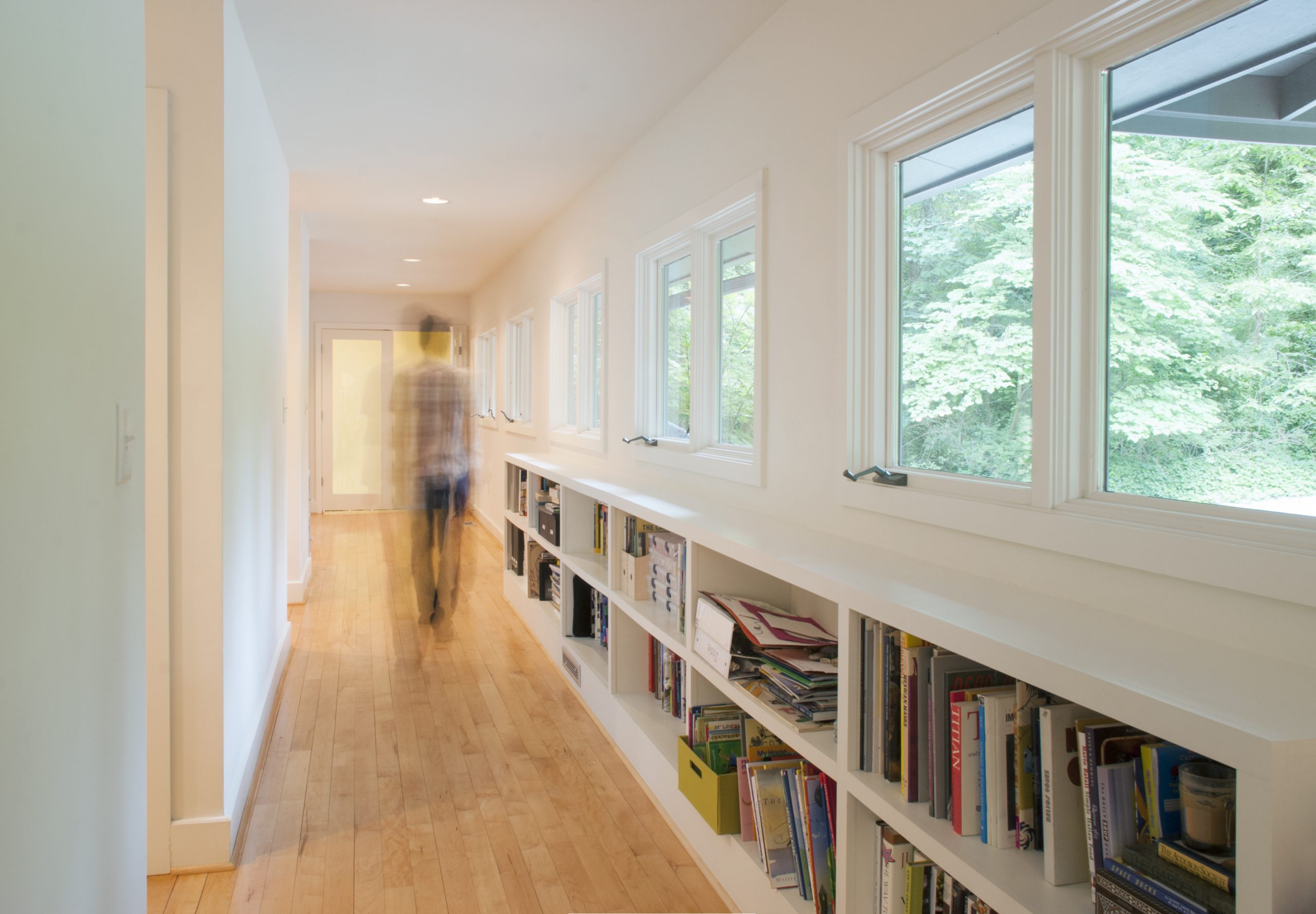 A person walks down a hallway lined with windows and low built-in bookshelves