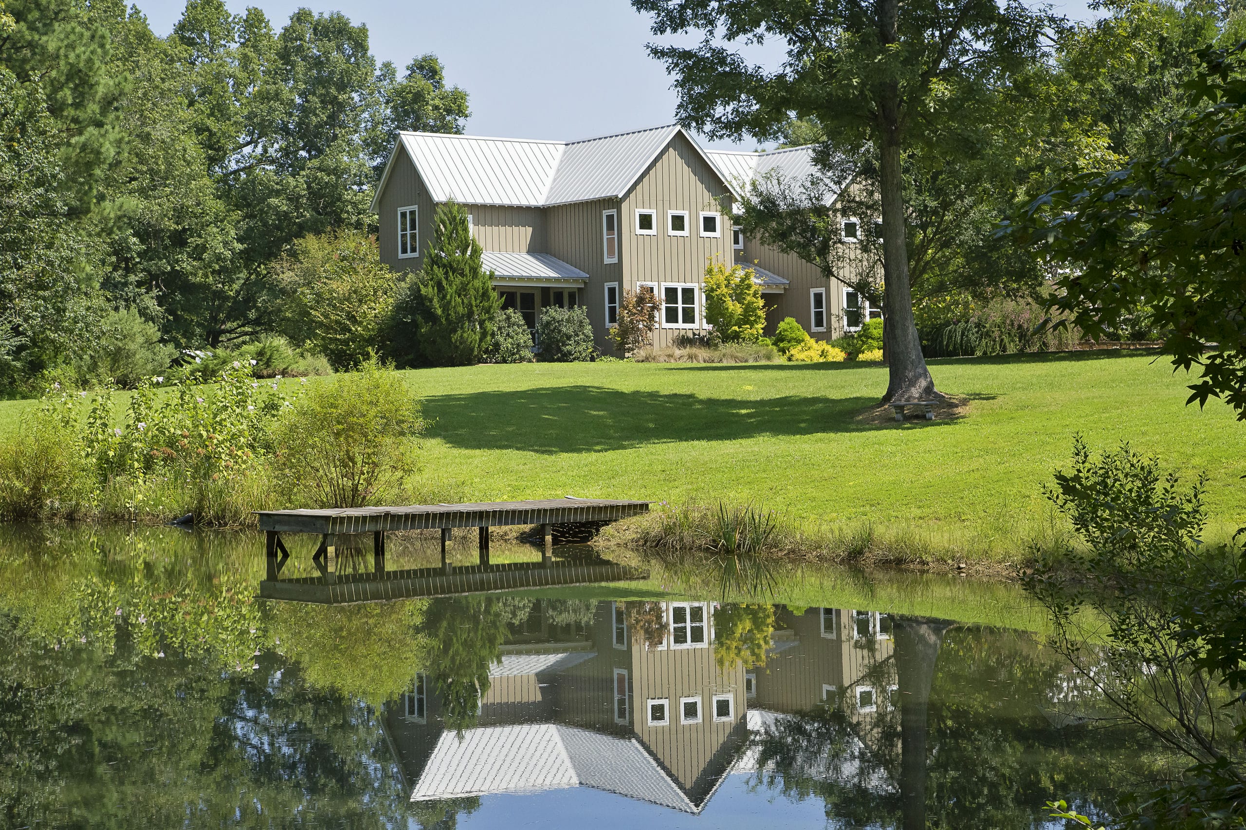 House with batten siding and gabled metal roof reflecting off a pond with wooden pier