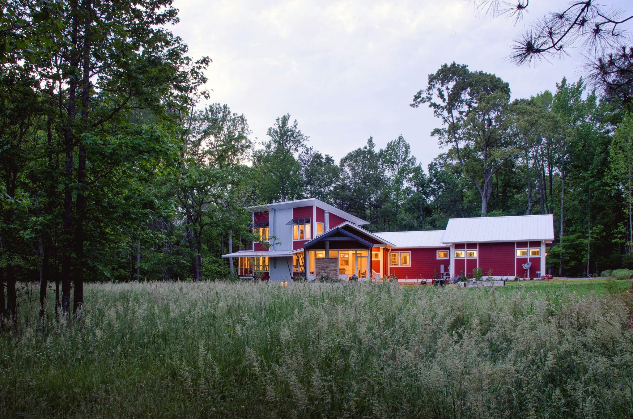 A view of the house at dusk, tucked away in tall grasses of a meadow. Red siding, white trim and standing seam metal roofs evoke a connection to the rural landscape.