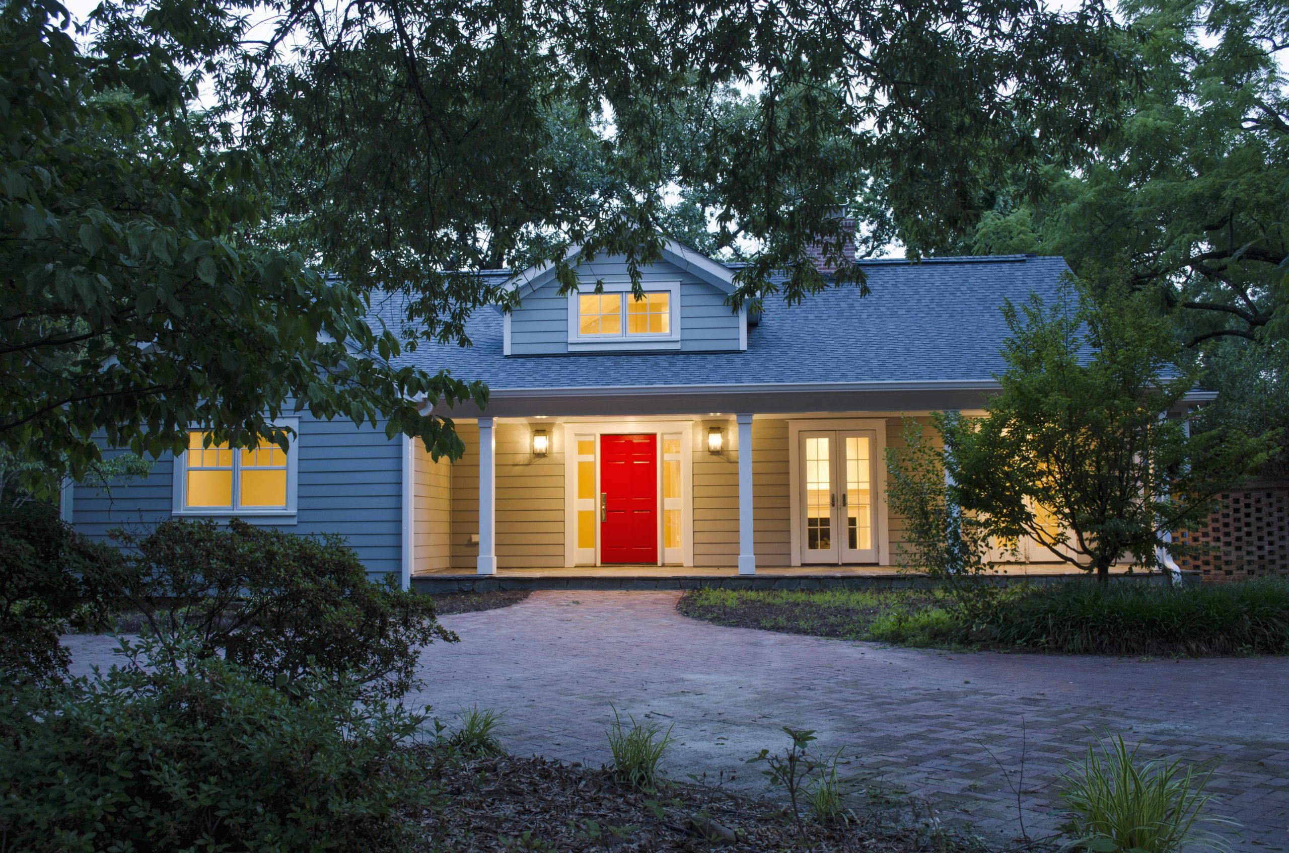 An exterior view of the house at dusk tucked away among the trees. The lights from within illuminate the front porch and red front door.