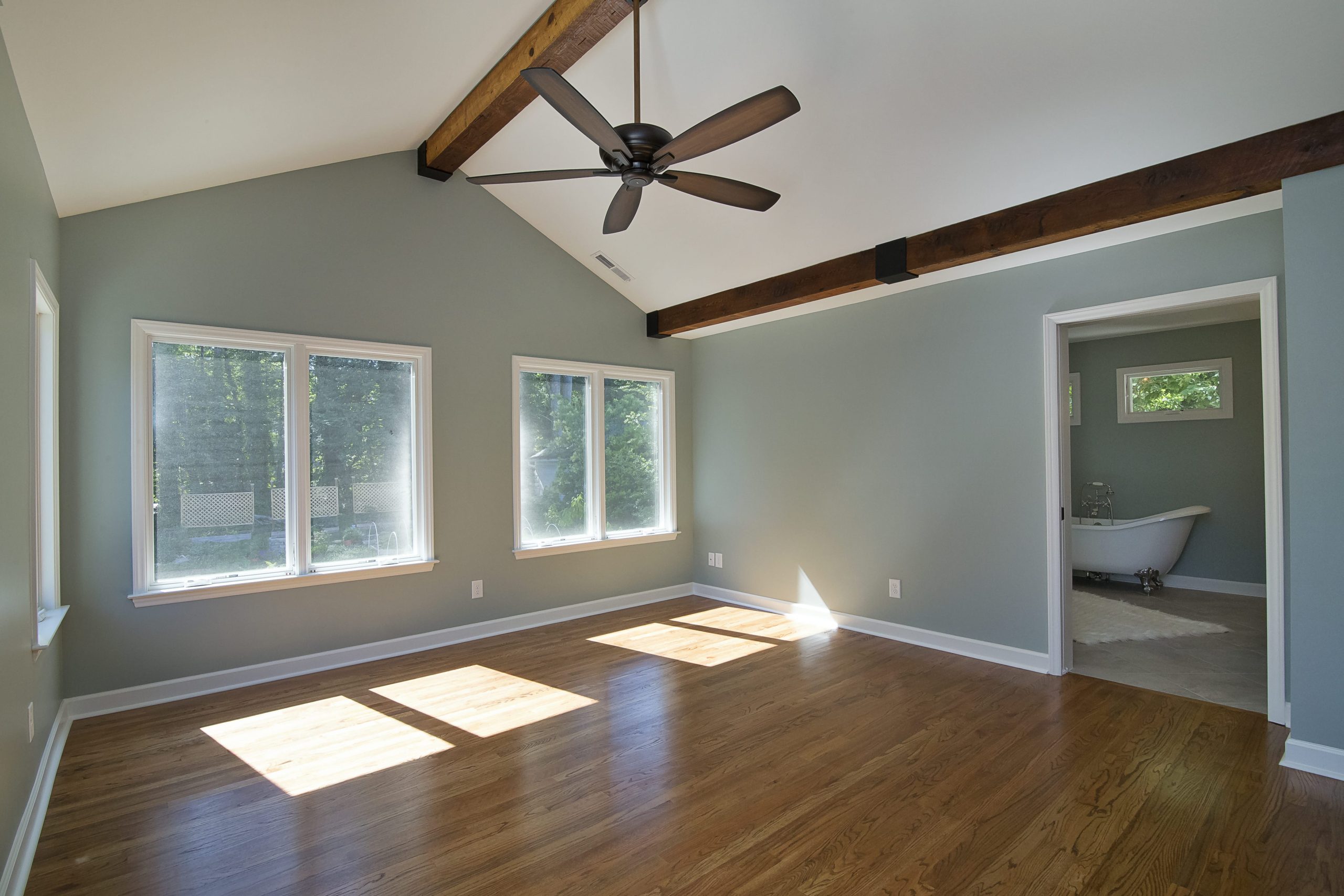 Primary suite with exposed wood beams and warm hardwood floors is illuminated by beams of sunlight through large windows