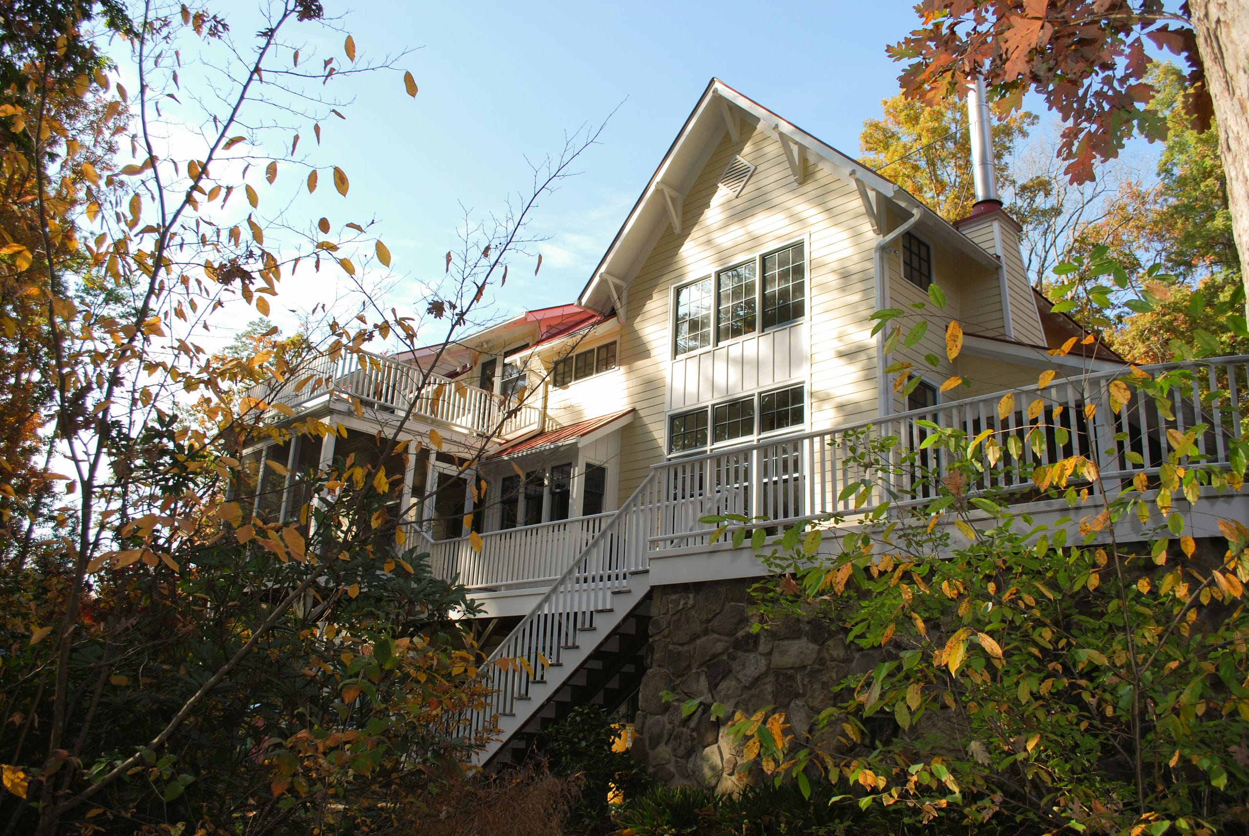 An exterior view of the house in autumn. Colorful leaves compliment the soft yellow siding, white window trim and unique red standing-seam metal roof.