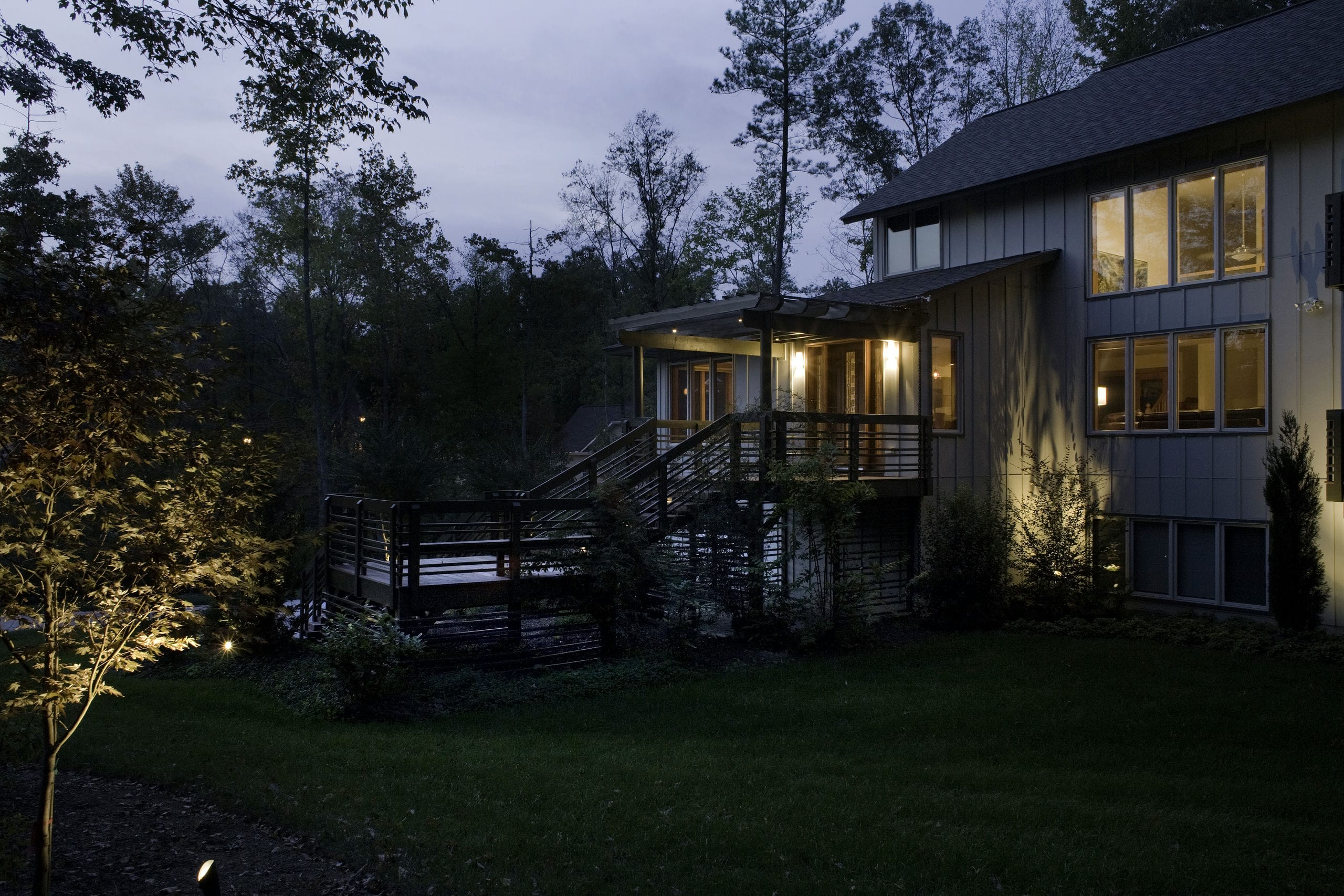 A nighttime shot showing exterior stairs leading up to trellis-covered entrance as landscape lighting and warm lighting from within the house illuminate the image.