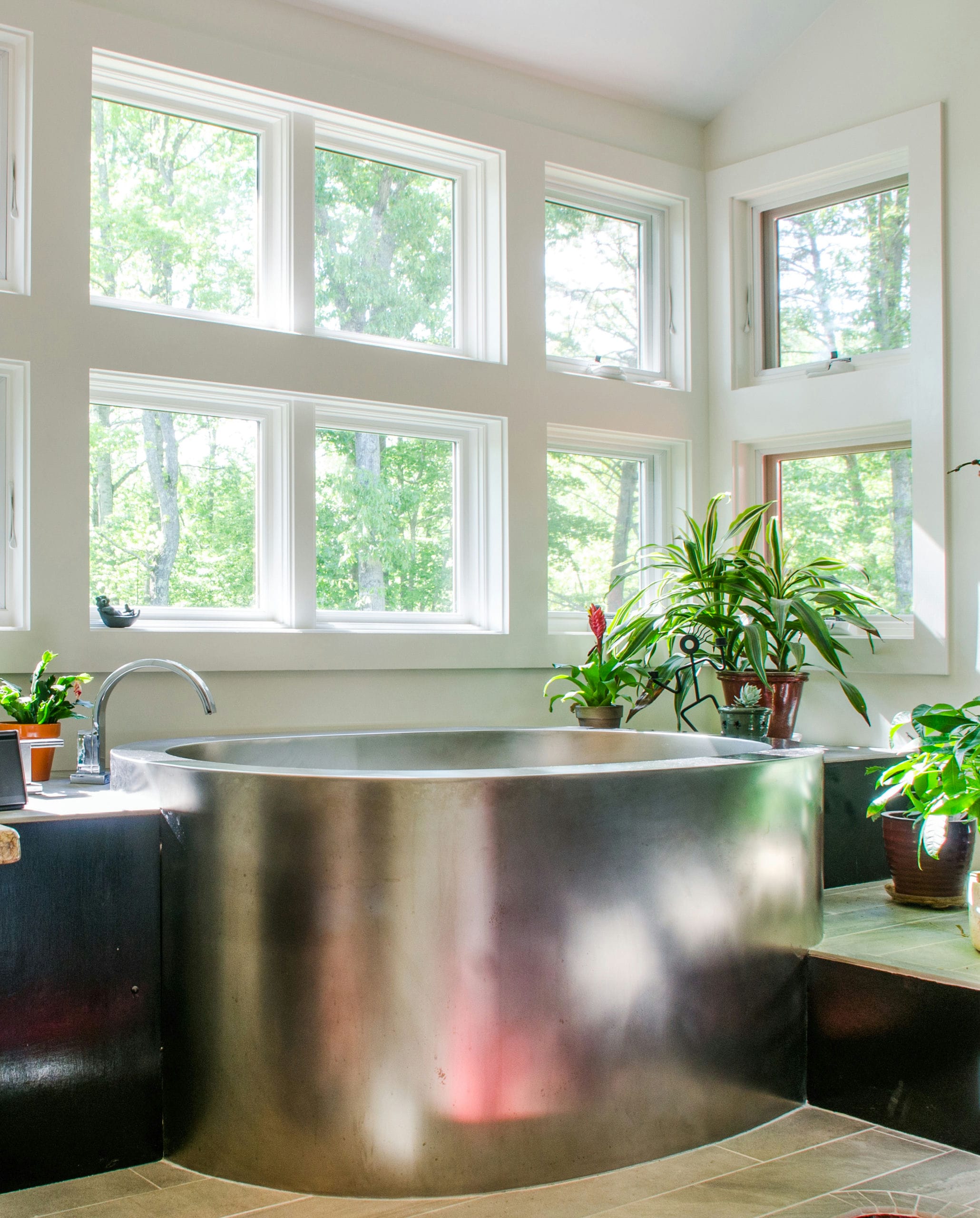 Sleek stainless steel Japanese soaking tub surrounded by plants and bay windows.