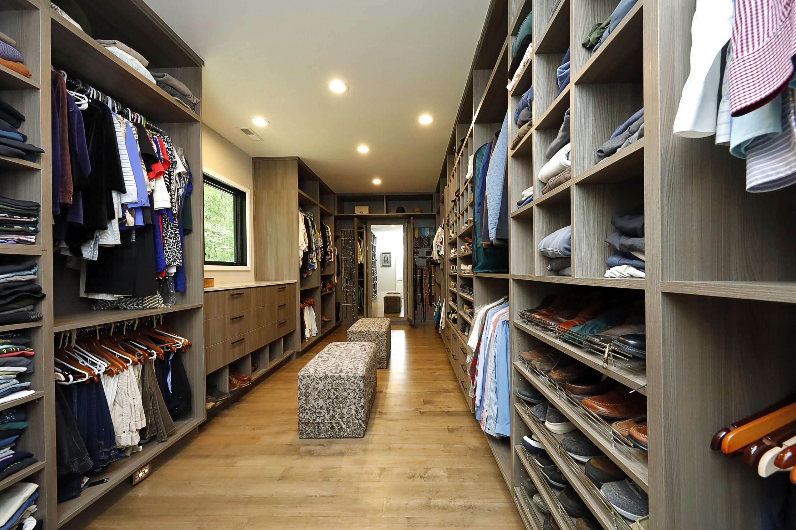 A large walk-in primary closet lined with built-in storage. A large central window illuminates a seating area.