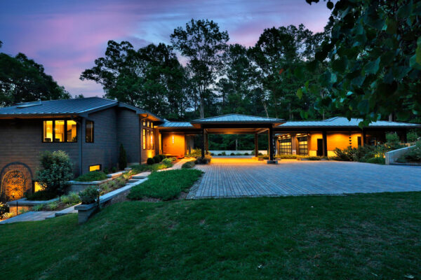 A view of the house at dusk showing the new garage, connected by a porte cochere, to the contemporary house. Warm lights illuminate the house from within.