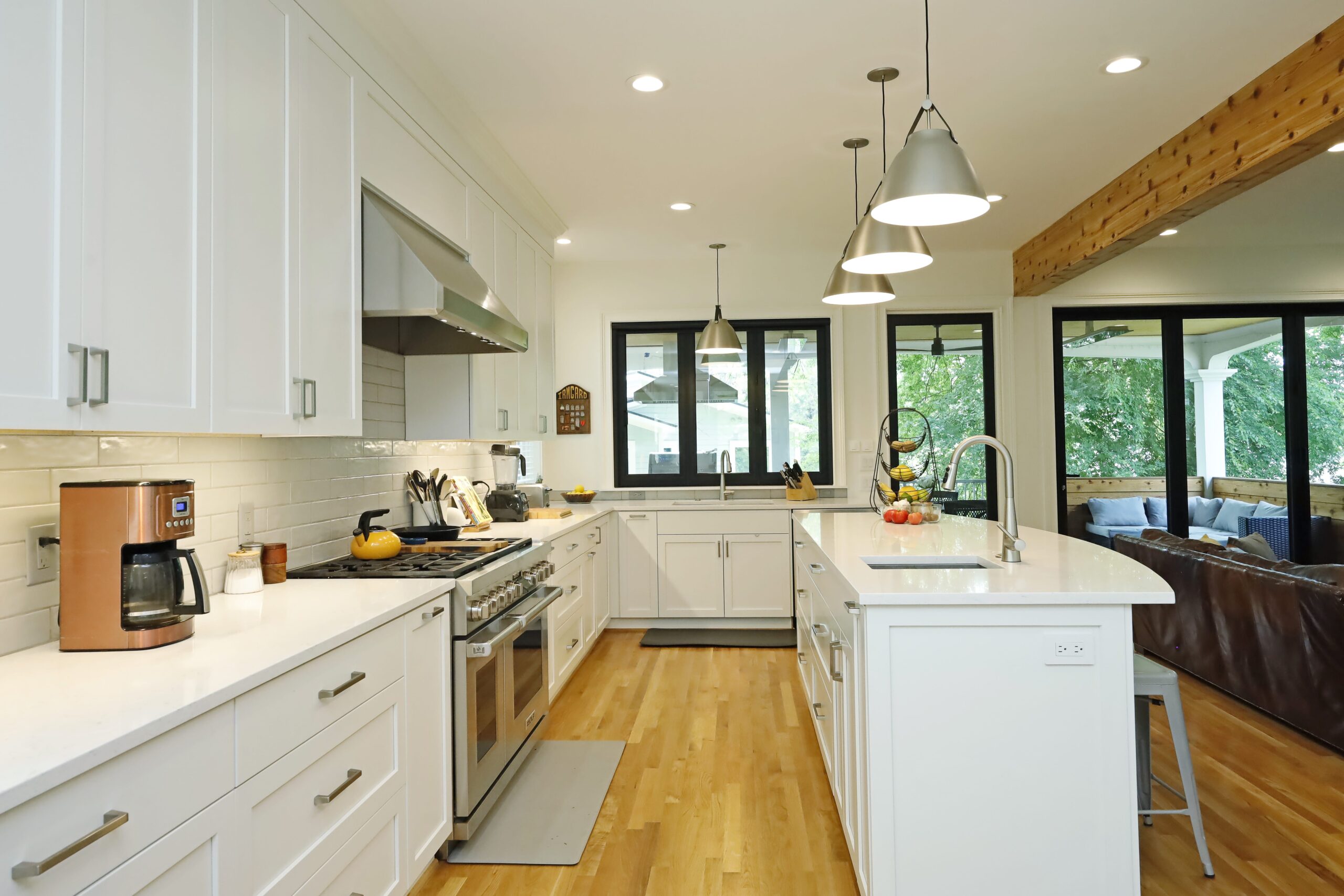 A view through the kitchen towards the covered deck highlights the generous kitchen prep space, sleek white counters and cabinets, and large accordions doors and windows onto the deck.
