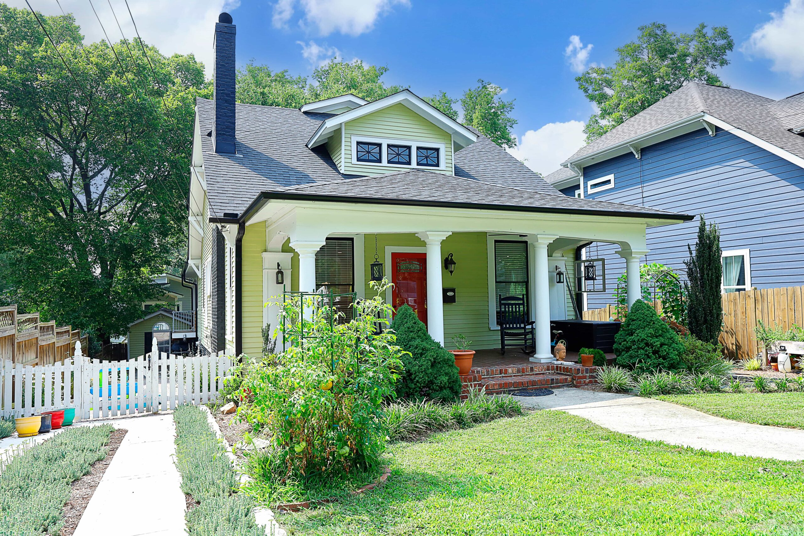 A view of the front of the bungalow style house. Green siding, a red door, a deep front porch, and a thriving garden add to the charm of this 1920s house.