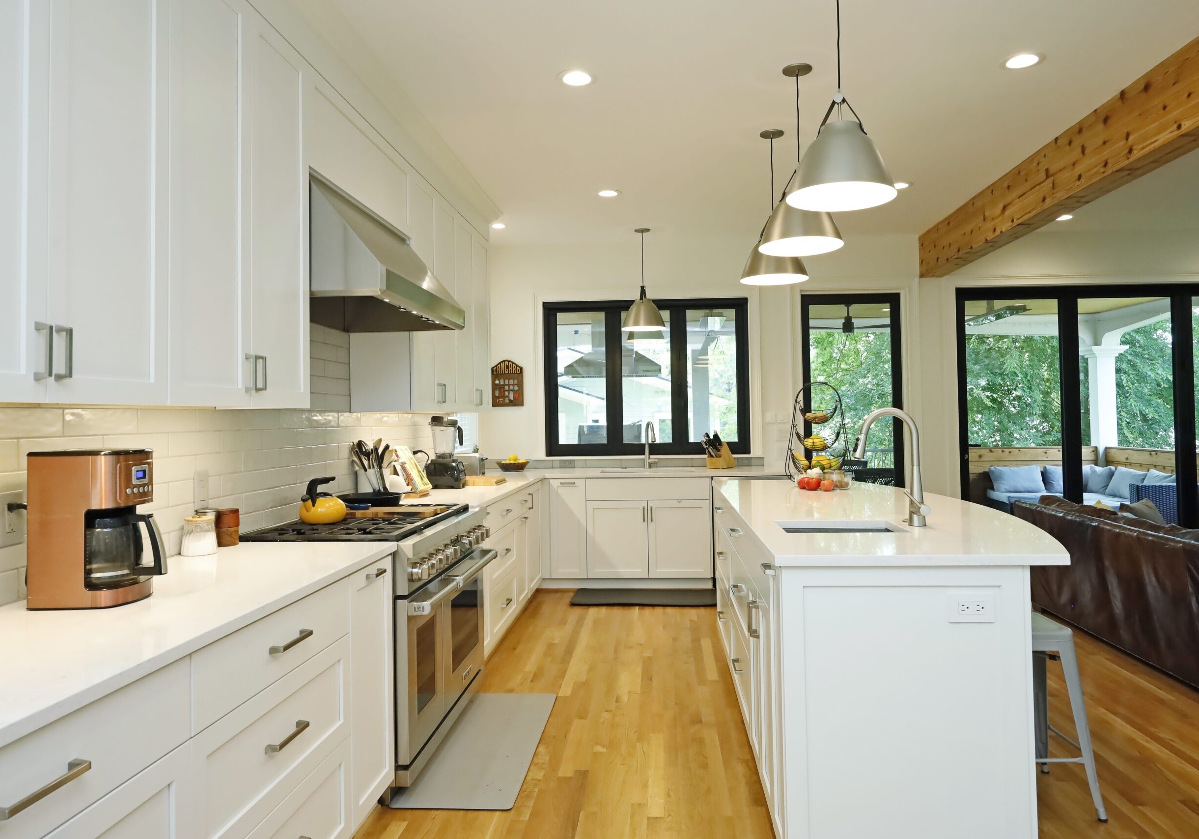 A view through the kitchen towards the covered deck highlights the generous kitchen prep space, sleek white counters and cabinets, and large accordions doors and windows onto the deck.